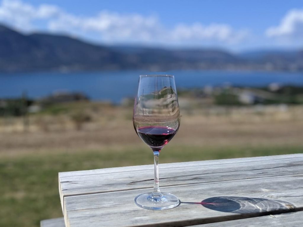Single wine glass on wooden picnic table at Origin Wines, containing tasting pour of Origin red wine. The background is Okanagan Lake.