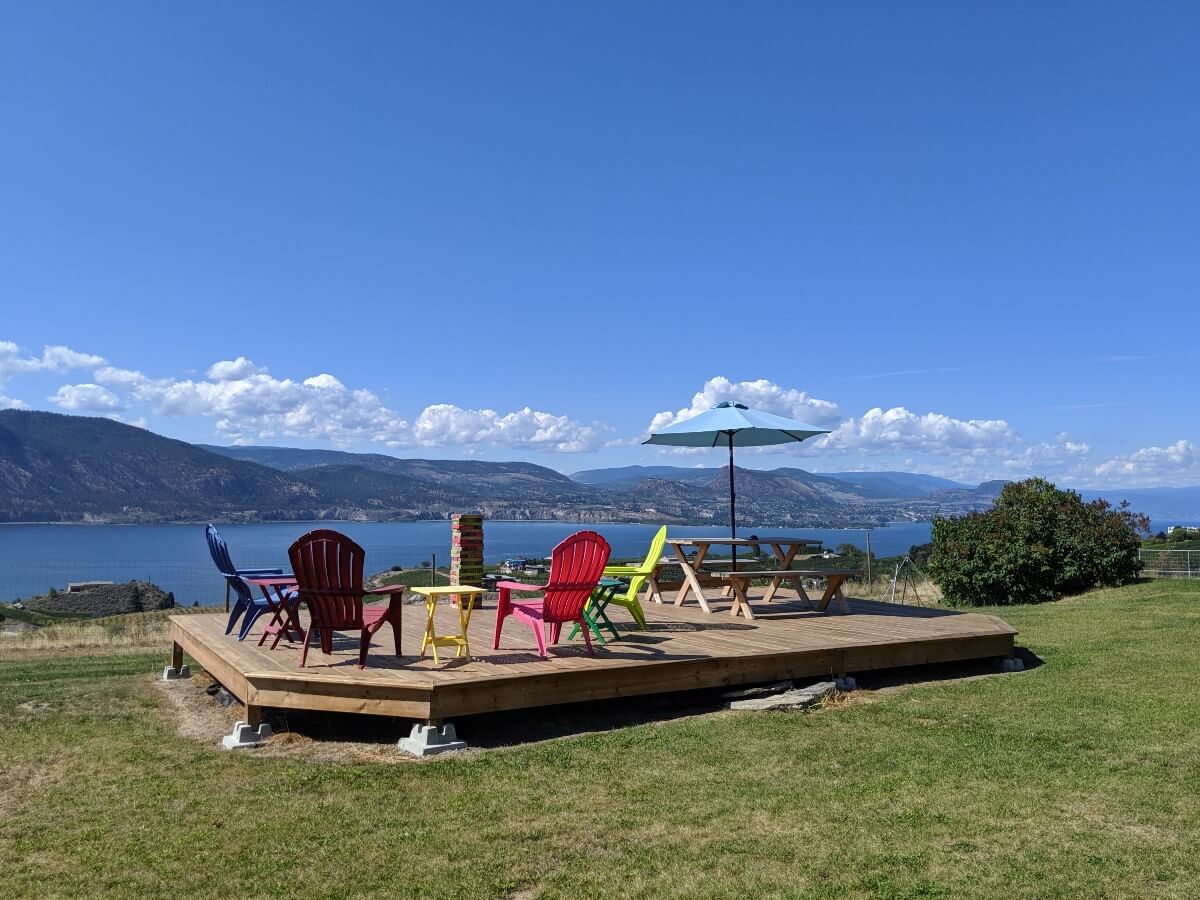 Outside patio seating at Origin Winery, with wooden deck, patio furniture and bench with umbrella, looking out to Okanagan Lake