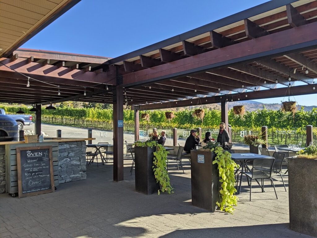Looking across paved area to outside patio at Upper Bench Winery, with 'the Oven' blackboard on left and seating on right
