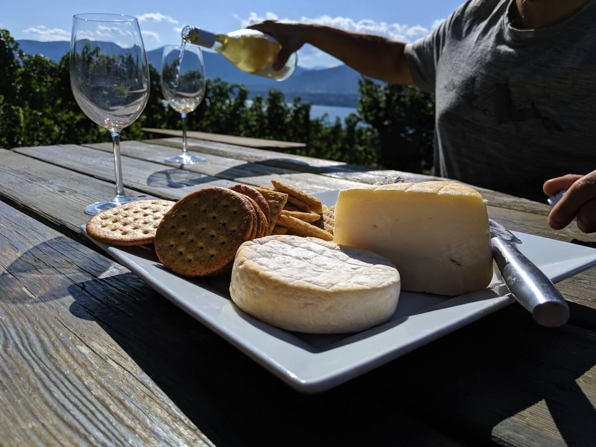 Side view of white square plate with cheese and crackers. In the background, a man is pouring wine into two wine glasses. There is a view of Okanagan Lake in the background