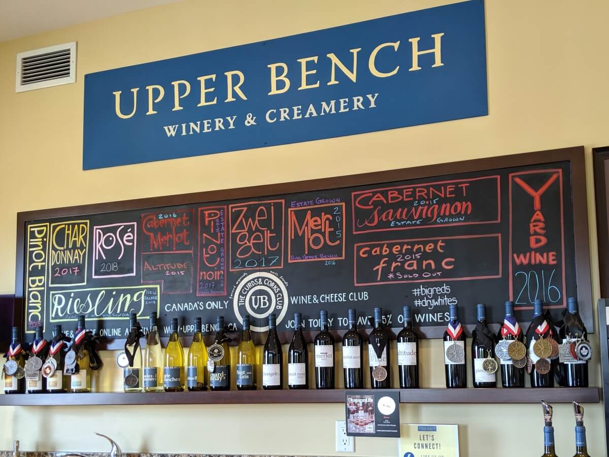 Wall of Upper Bench tasting room with logo at top and chalkboard underneath, with wine varietal names and wine bottles underneath