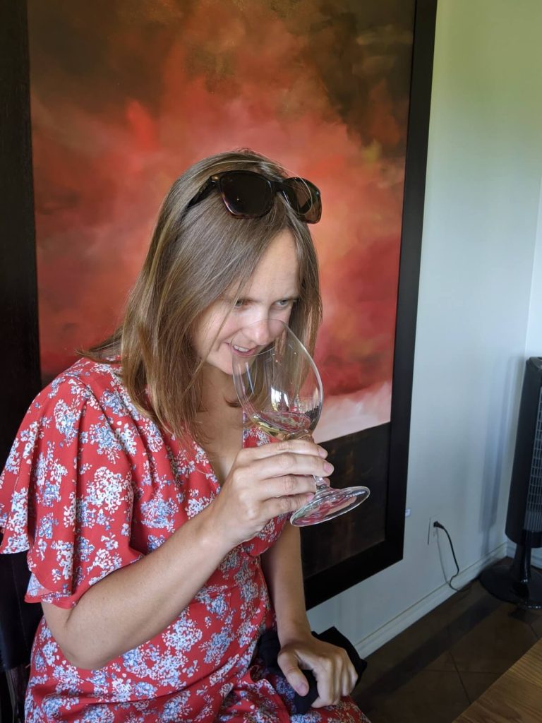 Women in red dress, in front of abstract orange painting, holding wine glass to her nose and smelling white wine.