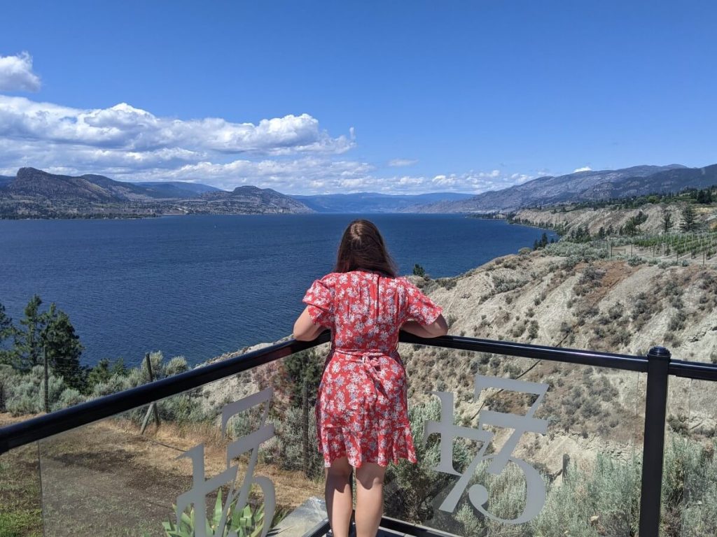 Back view of woman in red dress standing at edge of elevated Bench 1775 patio in front of Okanagan Lake and hills