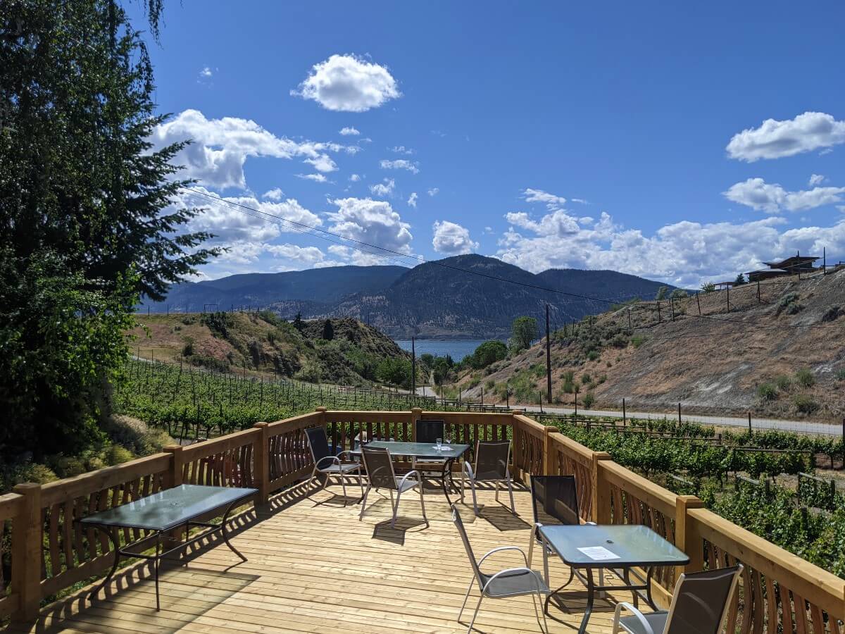 Looking over wooden patio above vineyard, with canyon and road view in background, slither of Okanagan Lake visible with forested mountain above
