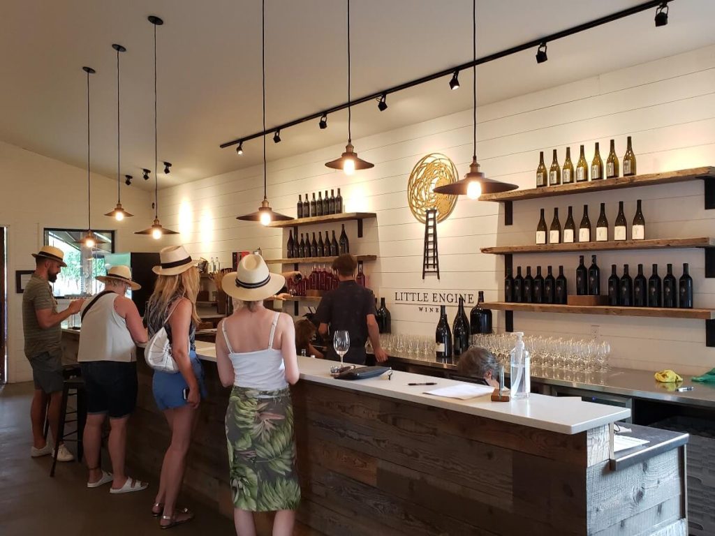 In the upscale winery tasting room at Little Engine, four people lined up in front of a fancy tasting bar. Behind the bar the Little Engine logo is on a wall made of white painted planks. On each side of the logo three shelves each showcasing a lineup of wines. 