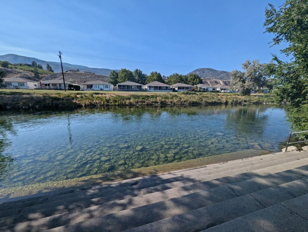 Looking down on concrete stairs leading into Penticton Channel, which featured calm and very clear river water. There are houses visible on the other side of the river