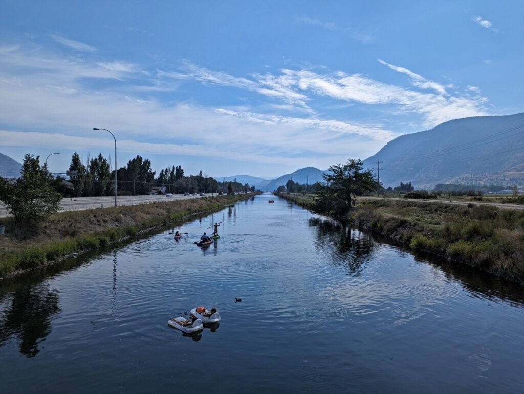 Bridge view looking down on groups of people sitting on inflatables and floating down Penticton Channel, which has sloped grass on both sides and a highway to the left