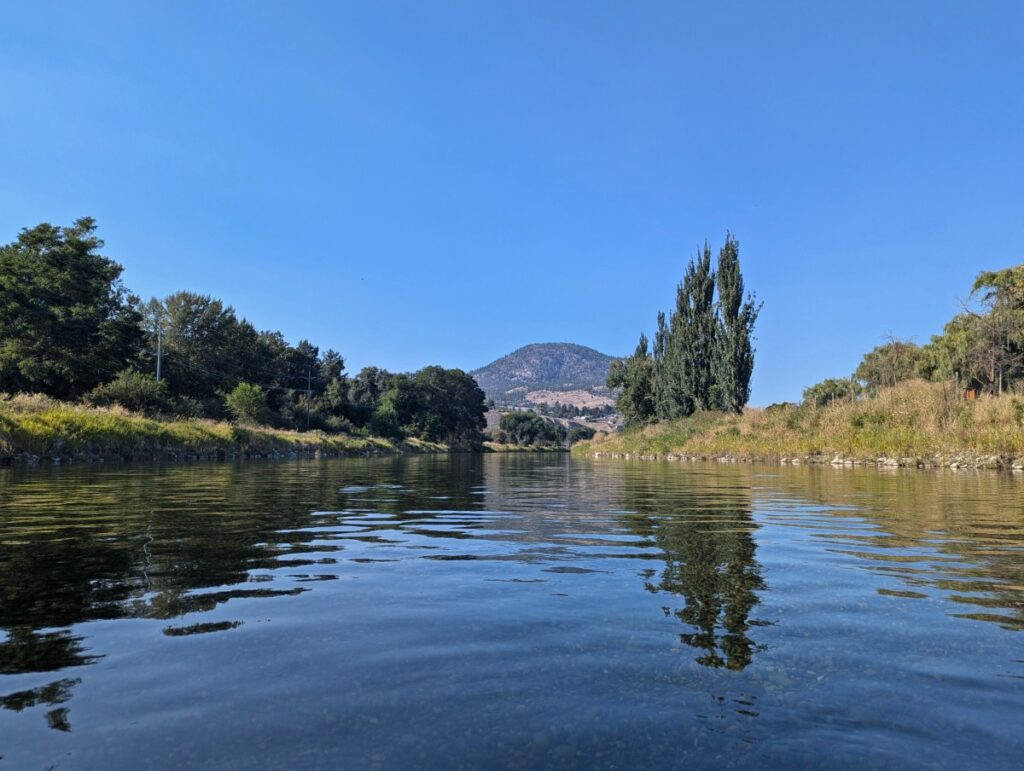 Water level view of floating the Penticton Channel, with calm river, grass and forest lining the edge, mountain in front