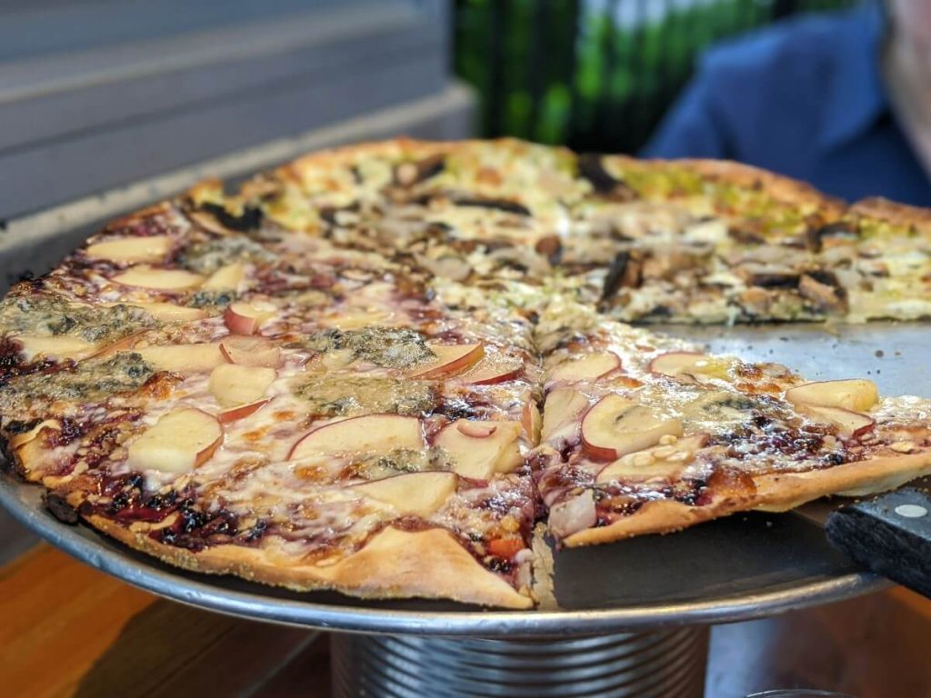 Side shot of large size pizza with one slice missing at Bad Tattoo. The pizza has sliced apples, jam and blue cheese melted on one half and mushroom and pesto on on the other half.