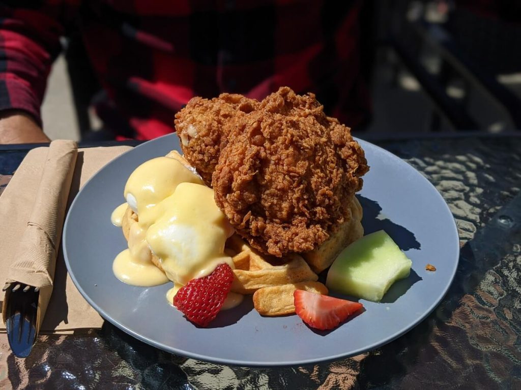 Close up of a blue plate at Loki's Garage featuring a waffle topped by some crispy southern chicken next to two eggs poached with hollandaise sauce. Next to the waffles are a couple pieces of strawberry and one slice of honeydew melon