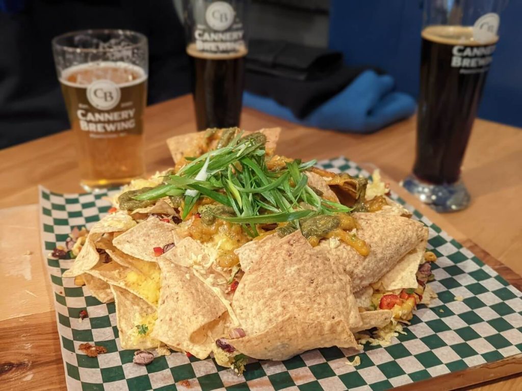 Mound of tortilla chips toped with green onions, melted cheese and avocado sauce on a checkered waxed paper. Blurred in the background, three Cannery branded pint of beers. 