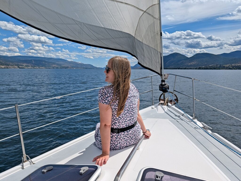 Back view of Gemma sat on the front of a yacht, with the sail visible as well as the calm lake and forested mountains