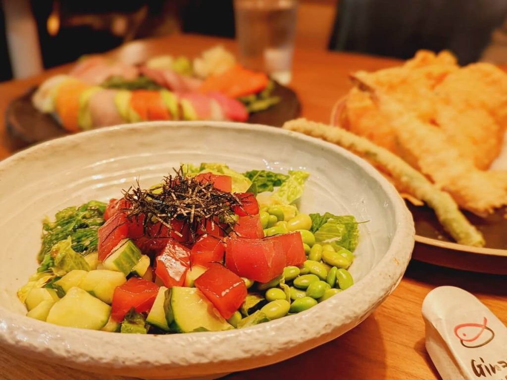Poke bowl at Ginza Sushi with cubes of tuna on greens and edamame toped with nori chiffonade. In the blurred background a plate of rainbow sushi roll and a plate of tempura vegetables 