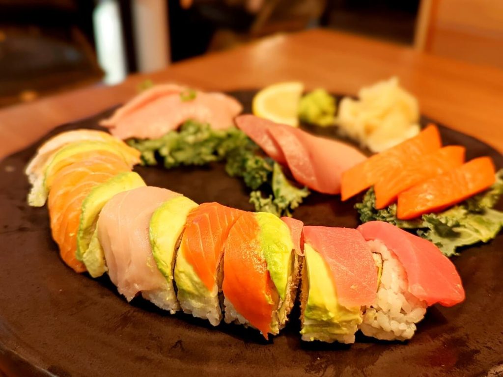 Colorful rainbow sushi roll at Ginza, consisting of tuna, avocado, salmon and white fish following the edge of the plate. On the same black plate 3 piece of each sashimi tuna, salmon, and white fish laid on a bed of kale. 