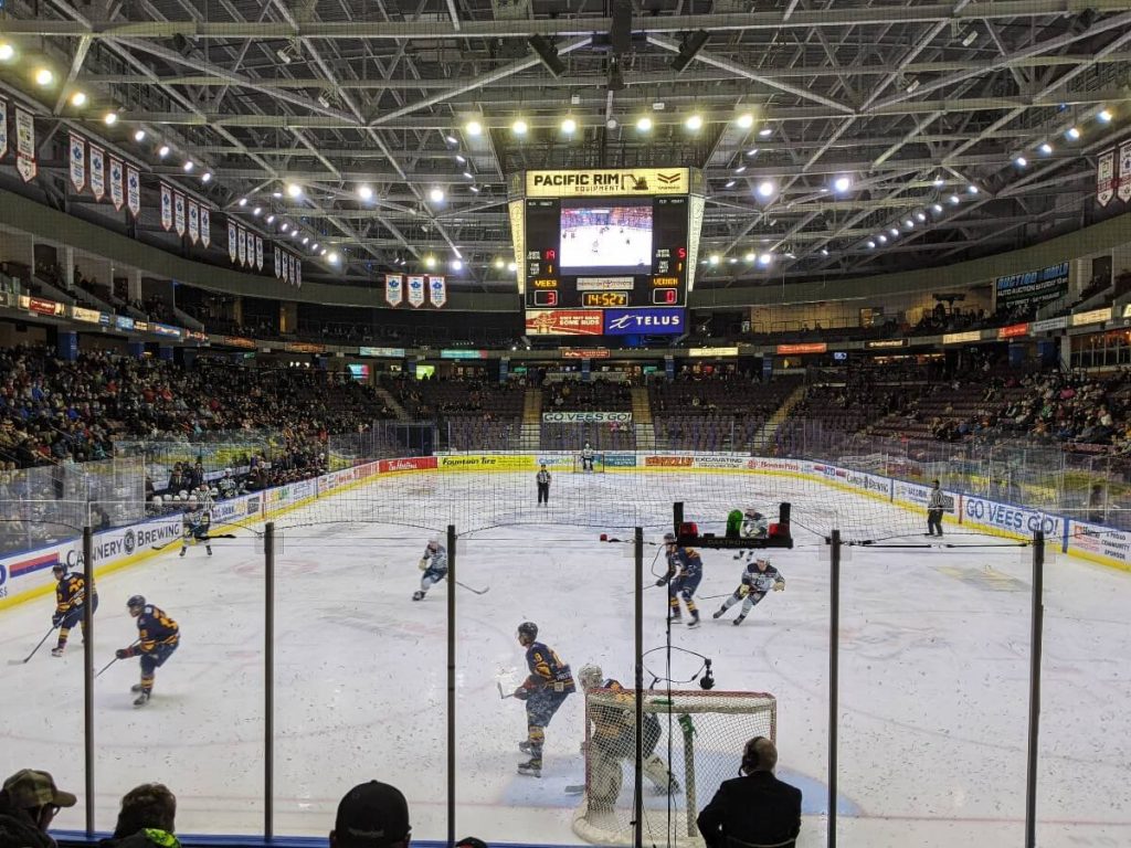 Ice hockey rink with players skating towards the puck. Above ice, a large score display three zero for the Penticton Vees. 