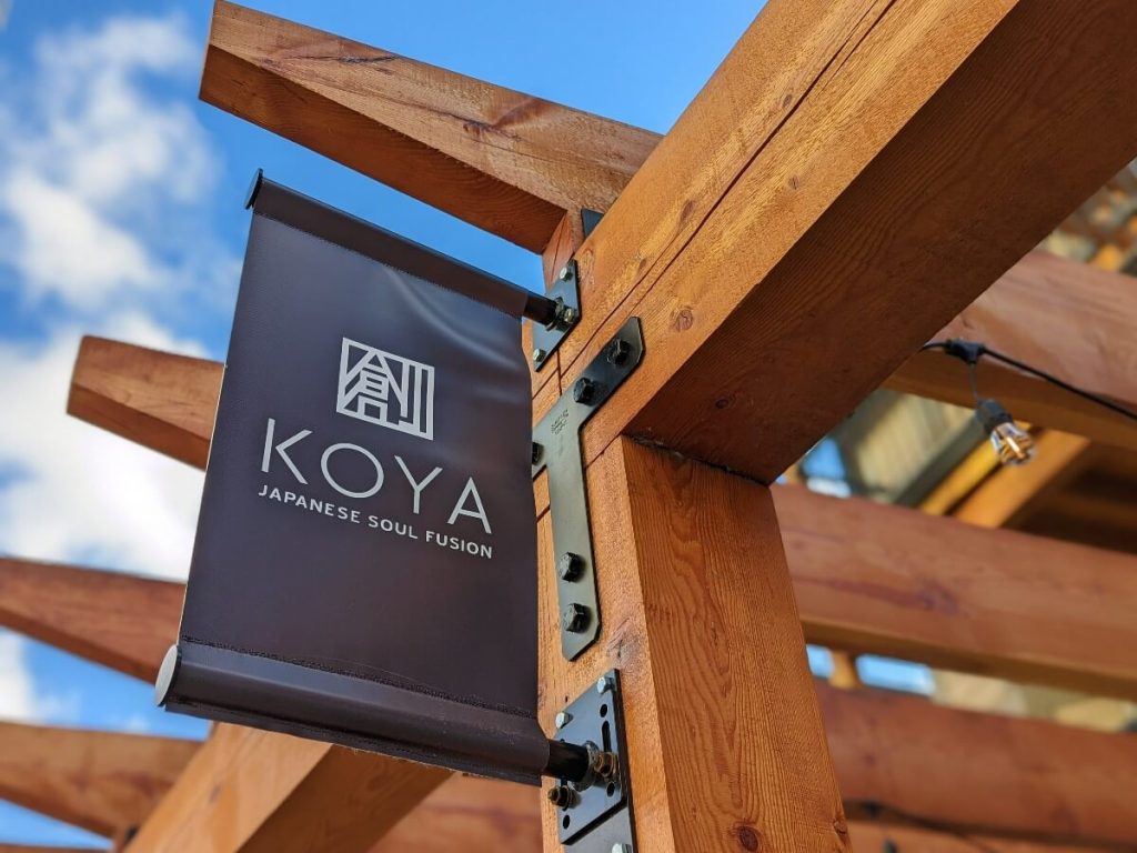 Black sign with Koya Japanese soul fusion in white writing hanging from wooden pergola beams.