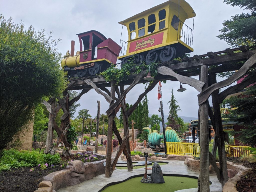 Red and yellow fake train and caboose with Loco Landing written on the caboose. The train is on a trestle passing over hole 16 of minigolf. In the background colorful yellow fence and a green Ogopogo statue