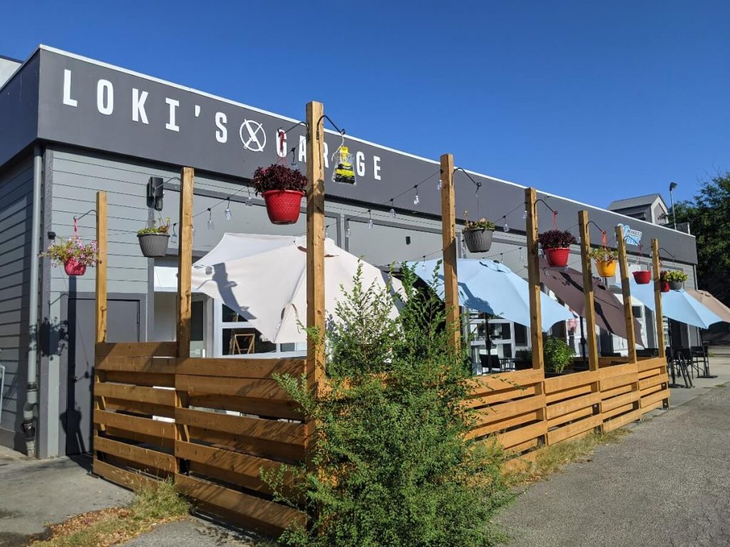 Loki's Garage sign painted on the upper part of one story building. The gray building has a wooden plank fence surrounding the patio with flower pots hanging from each fence post. The patio has five umbrellas shading the tables from the sun. 