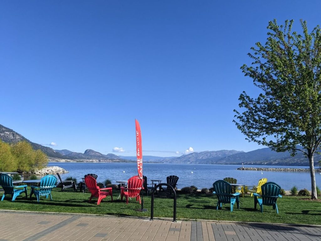 Back view of colourful Prague Café patio chairs on grassy area in front of sandy Marina Way Beach in Penticton