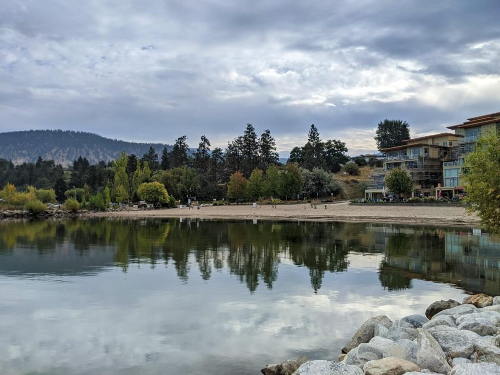 View of the sandy beach of Penticton Marina beach with reflection of trees in the water.