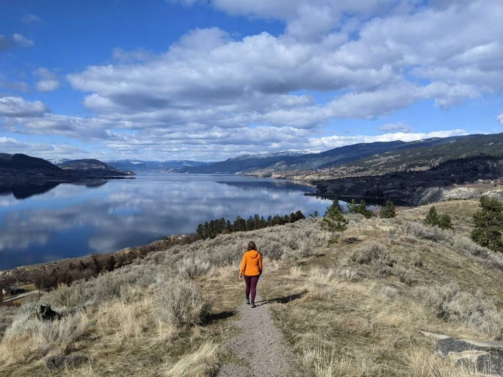 View of Okanagan Lake from Munson Mountain lookout trail with woman in orange jacket walking away from camera