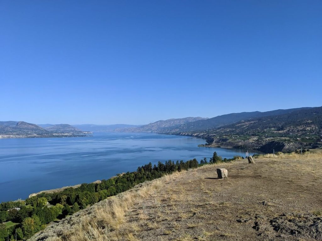 Flat summit area on Munson Mountain, with two of the stones visible. Okanagan Lake is seen below peak to the left, with bluffs and vineyards behind on the right
