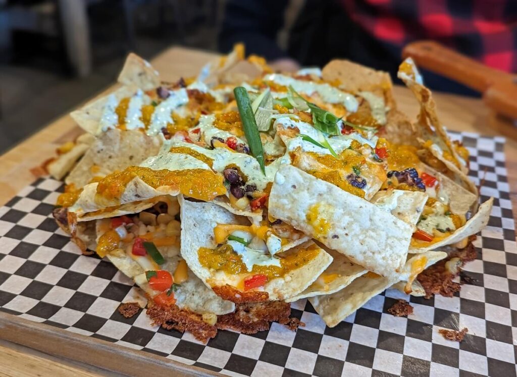 Mound of tortilla chips toped with green onions, melted cheese and avocado sauce on a checkered waxed paper. 