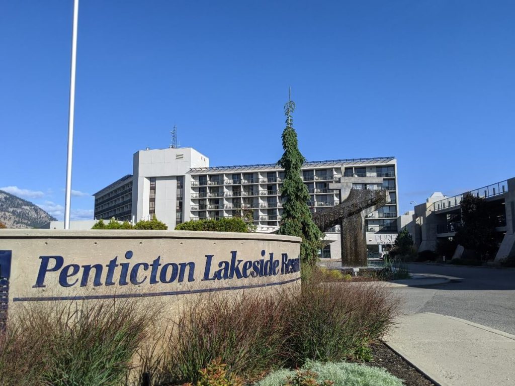 Penticton Lakeside Resort concrete corner sign with logo. In the background, a stone sculpture fountain and the gray hotel building.