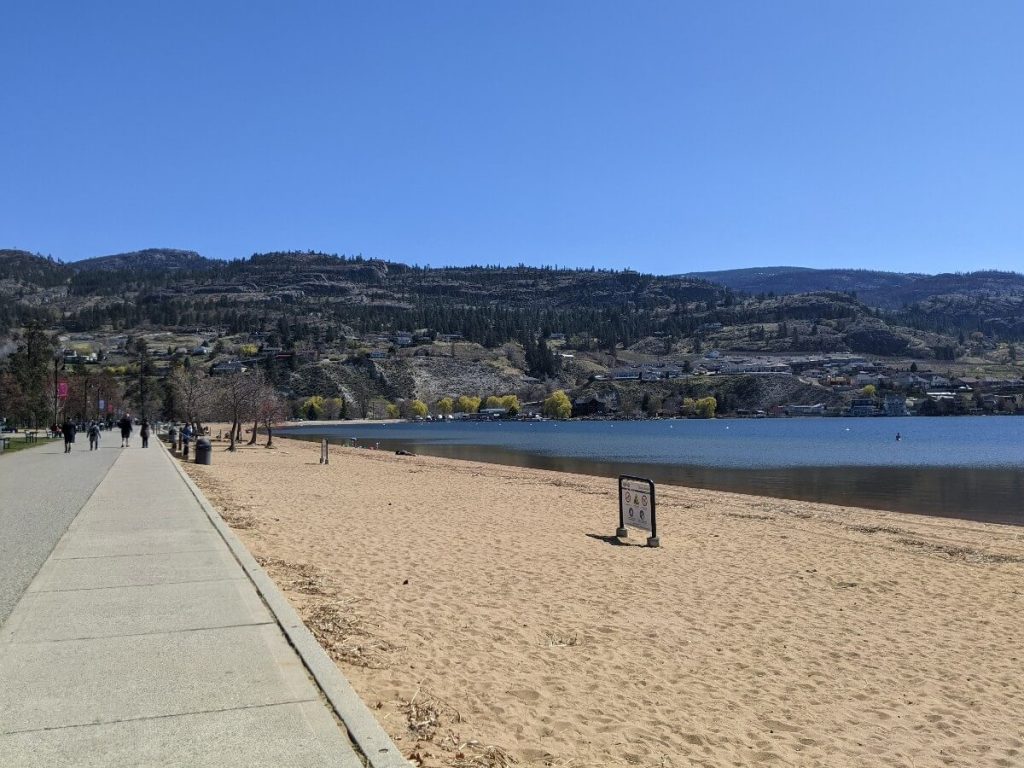 People walking on the elevated concrete promenade at Skaha Beach, next to golden sand beach and calm Skaha Lake