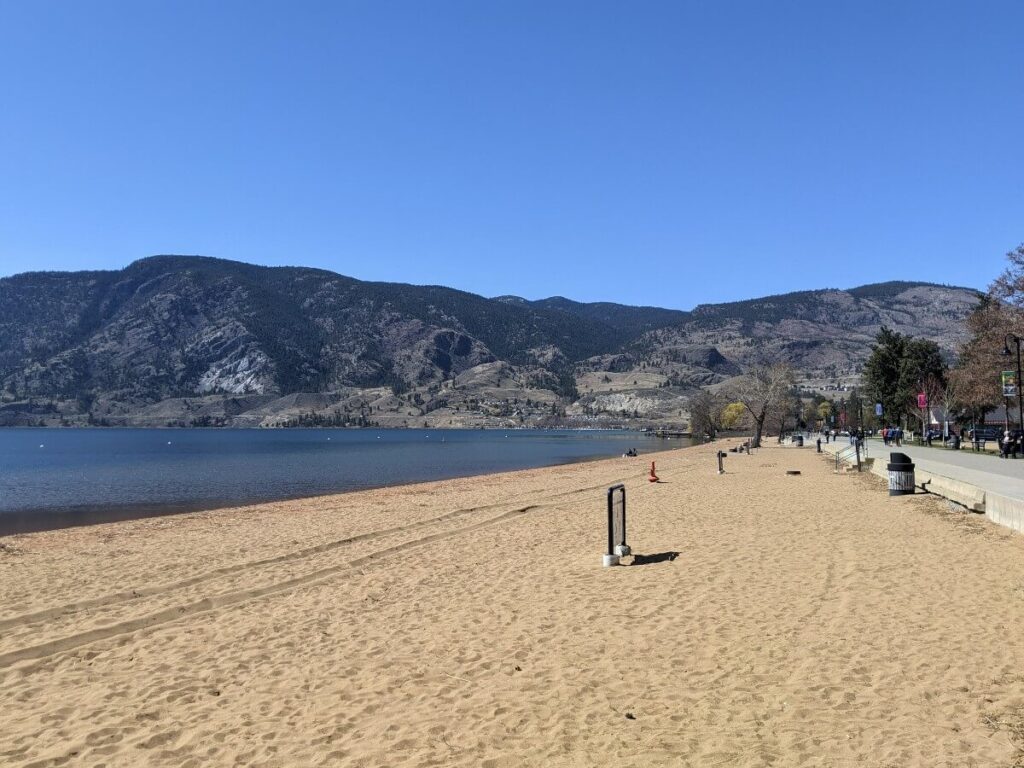 View of Skaha Lake Beach with hills in the distance and people walking on lake shore drive walking path.