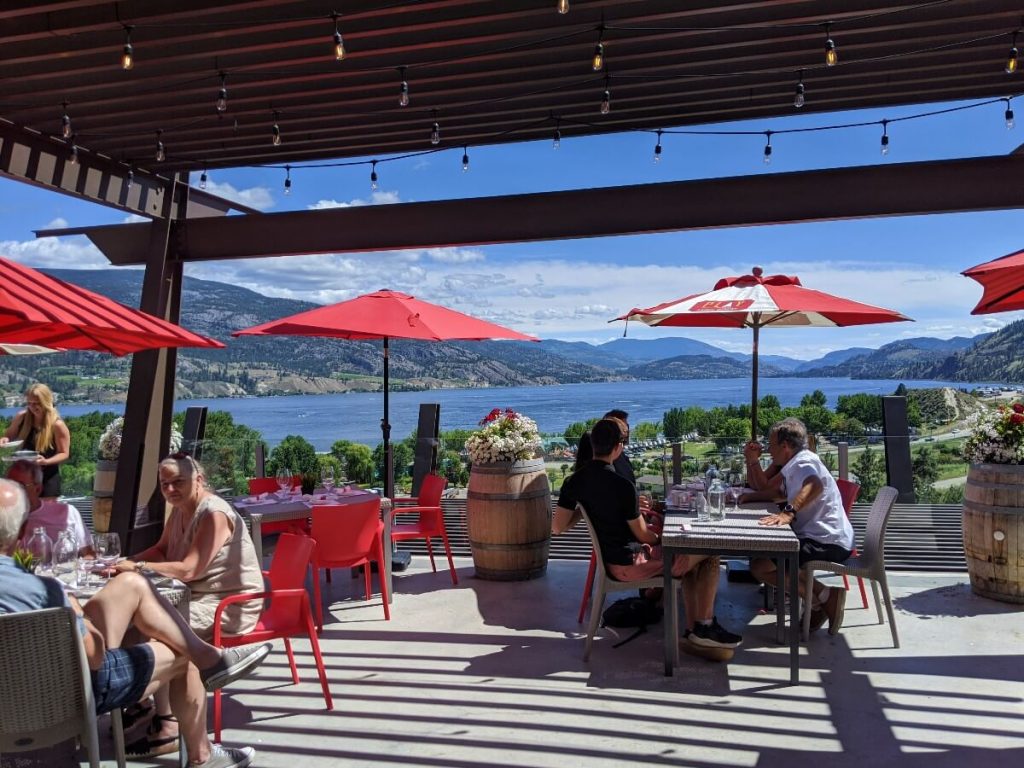 Patio at Play Winery featuring tables and red chairs on concrete surface with red umbrellas. In the background is a beautiful view of Skaha Lake, surrounded by mountains