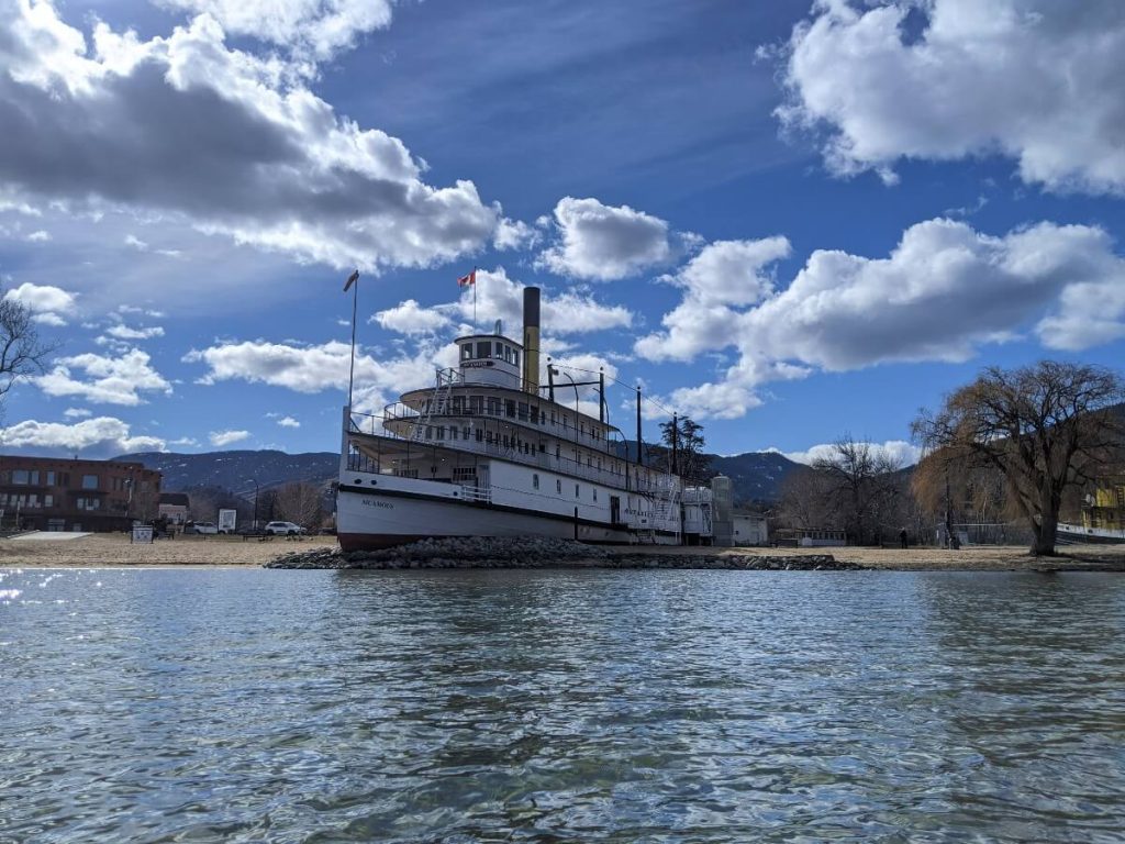 Boat view of large white SS Sicamous paddlewheeler on land, surrounded by rocks, with blue sky and fluffy clouds above
