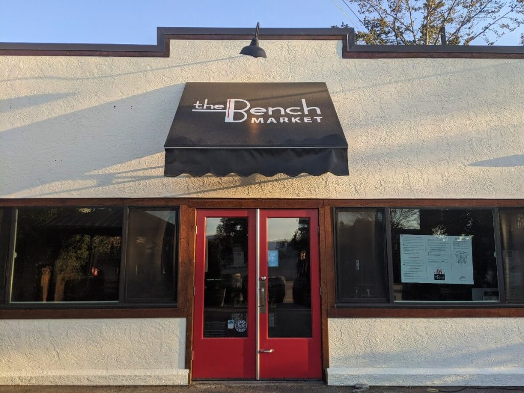 The Bench Market building with large windows and red doors under black awning above them. 'The Bench Market' in white letters is written on the awning.