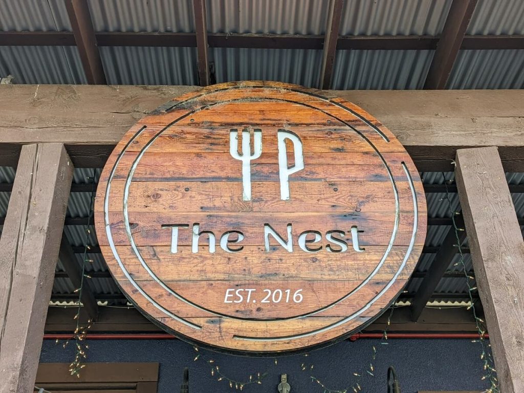 Close up of wooden round sign with The Nest written on it. A fork and a knife, carved into the wooden sign, stand parallel to each other  above the name on sign.