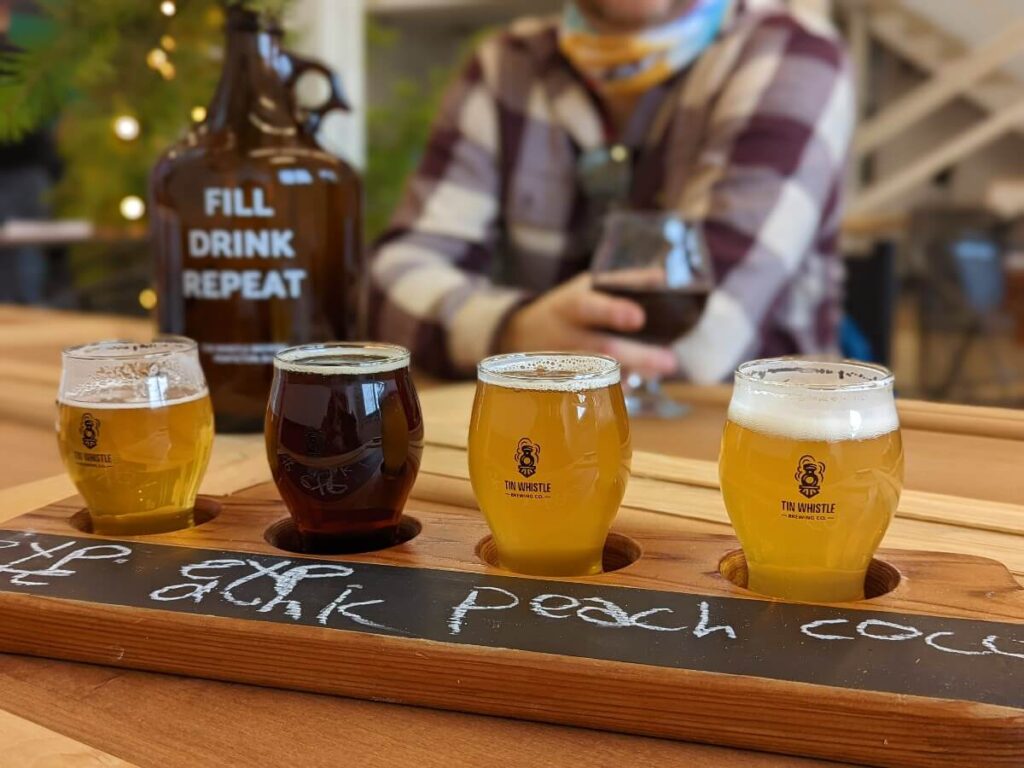 Tin Whistle Brewing tasting flight of 4 beers lined up in its holding tray and description of beer written in white chalk on black line of tasting tray. Beer growler and person holding beer glass in blurred background. 