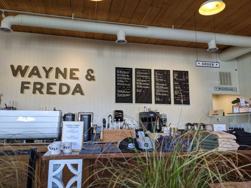 View of Wayne and Freda serving counter and display of branded merchandize on a long wooden table seen trough decorative grass.  Four black black boards display the menu next to the Wayne and Freda logo painted on wall. 