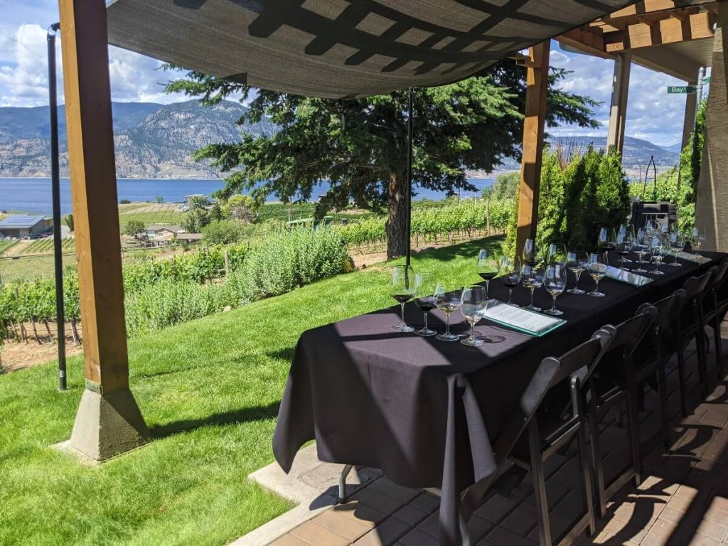 Black table clothed table with chairs and four lined up wine glasses in front, looking out to vineyards and Okanagan Lake