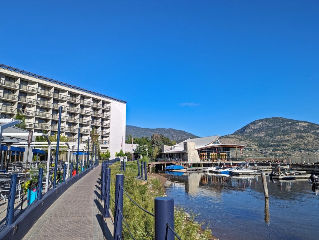 Looking along Okanagan lake paved walkway with Barking Parrot patio on left, Lakeside Resort hotel above and marina on right