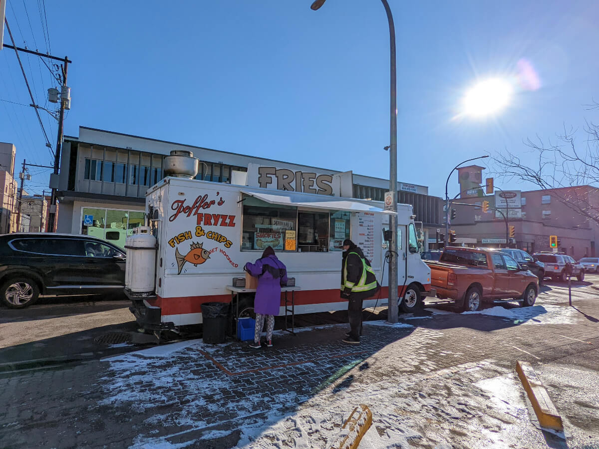 A white truck adorned with Jeffer's Fryzz signs is parked by the side of a slightly snowy street in downtown Penticton on a sunny day. 