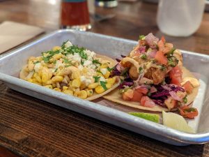 Two tacos on a silver colored tray at Neighbourhood Brewing. One taco with fried fish, diced tomato and red onion. The other is street yellow corn toped with cheese and cilantro.