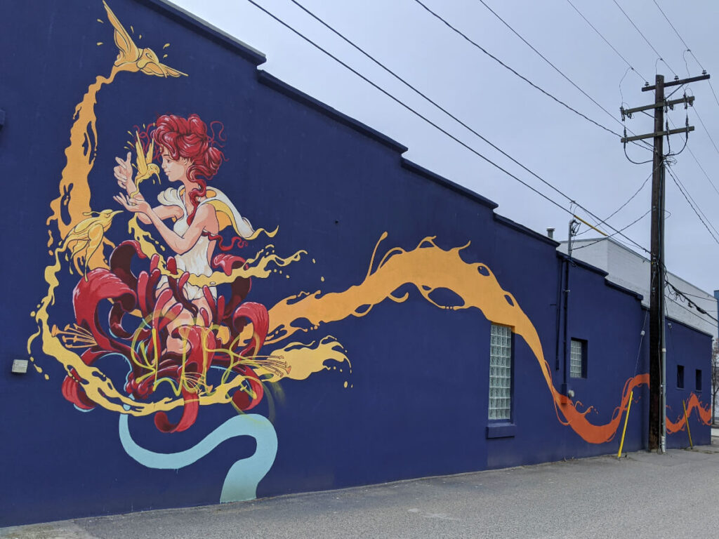 Side of building in downtown Penticton with brightly coloured street art, blue design with woman in orange/red