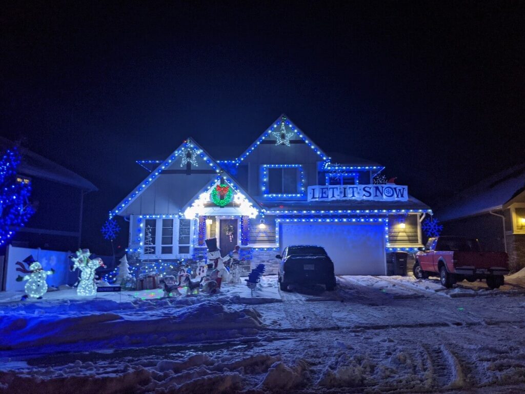 Roadside view of brightly lit house on Lawrence Avenue, with blue theme. There are large snowmen figures on the lawn and a Let It Snow sign above the garage