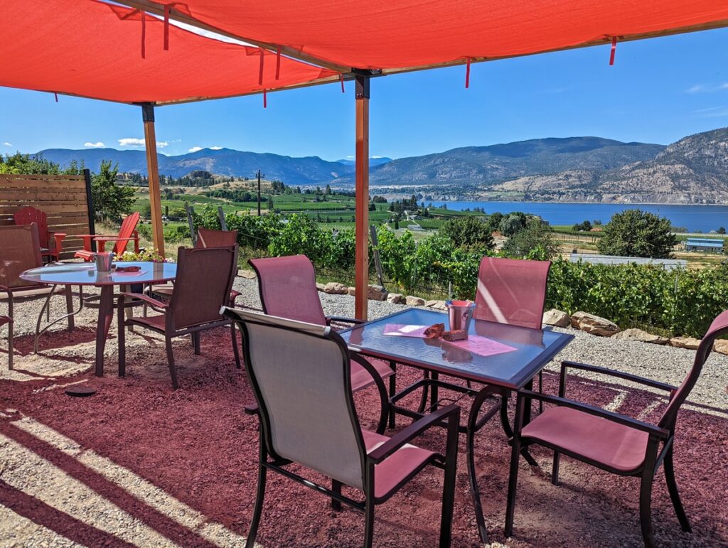 Two sets of tables and four chairs on shaded patio at Chain Reaction Winery with lake and vineyard views in background