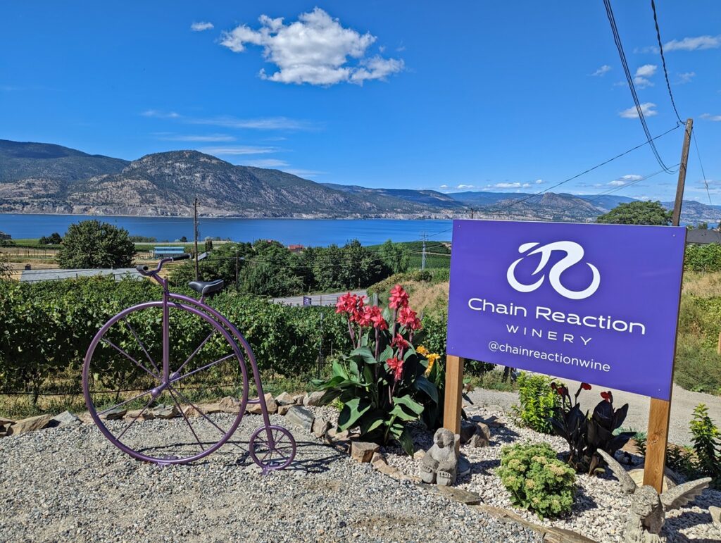 Purple Chain Reaction Winery sign next to old penny farthing style bike, with views of Okanagan Lake behind