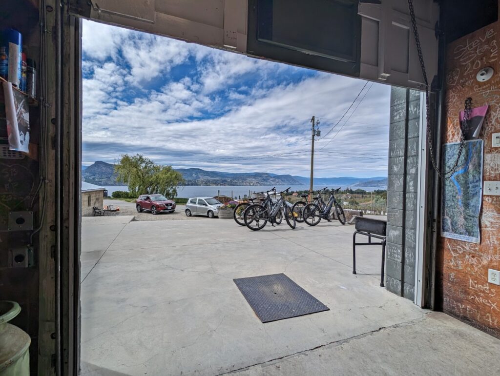 Looking out through the large open door at Van Westen Vineyards to a full bike rack with parking behind. Okanagan Lake is visible in the background