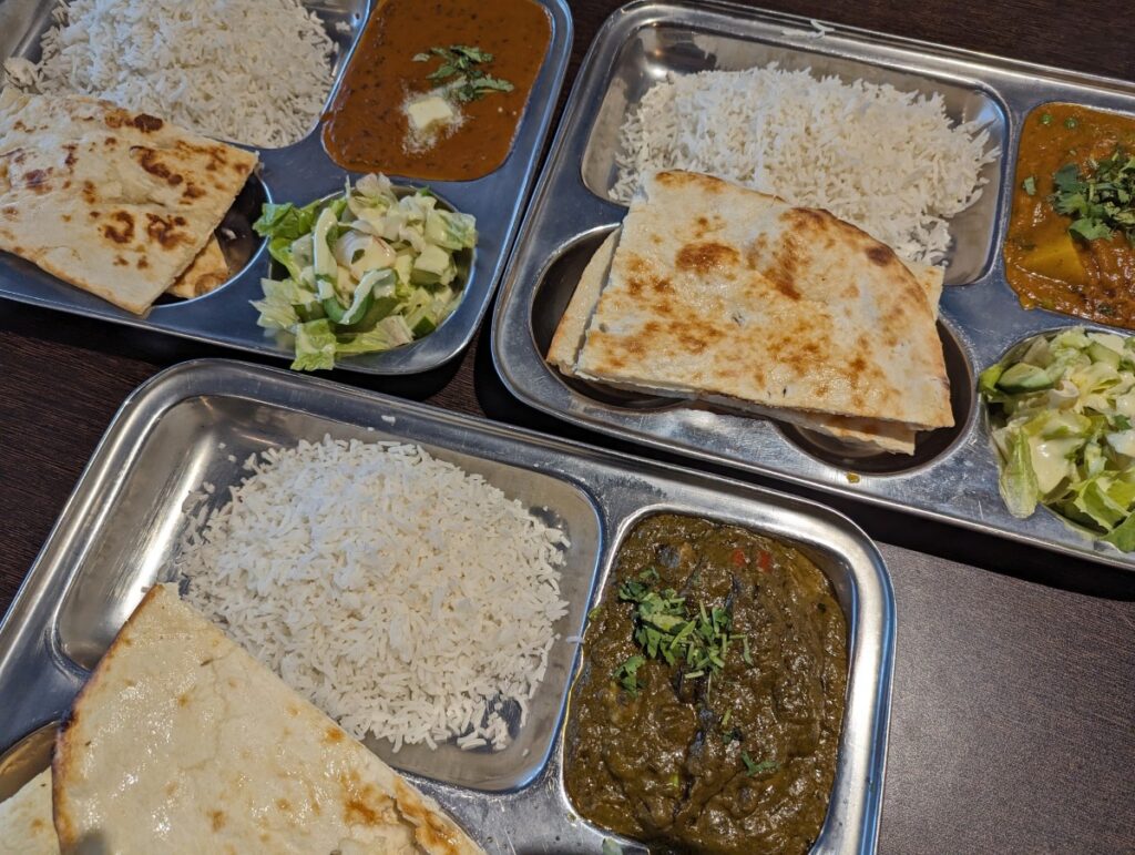 Overhead shot of three metal combination platter plates at Ashoka Indian Cuisine in Penticton featuring curry, rice, salad and naan bread