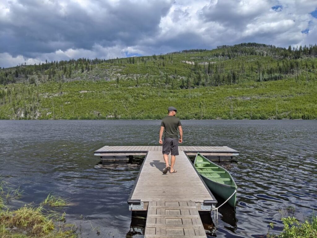 Back view of man walking on wooden floating dock on Chute Lake, with canoe on right land side. The lake is calm and surrounded by shrubs and trees