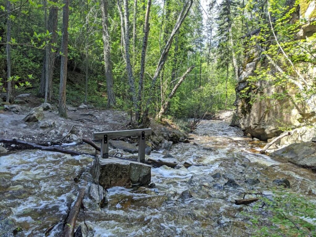 View of Naramata Creek Falls crossing, with remains of wooden bridge. Backdrop of trees and cliff to right