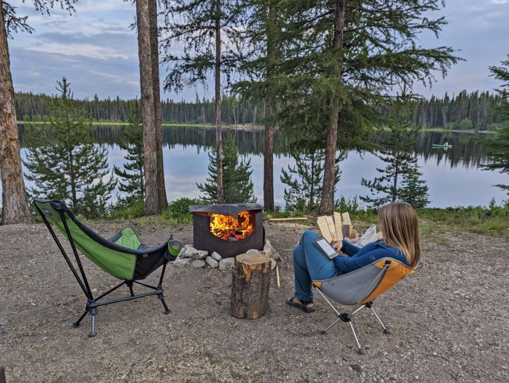Back view of woman sat in chair reading e-book in front of lit metal fire pit. There is an empty chair on left. A reflective lake is visible in the background