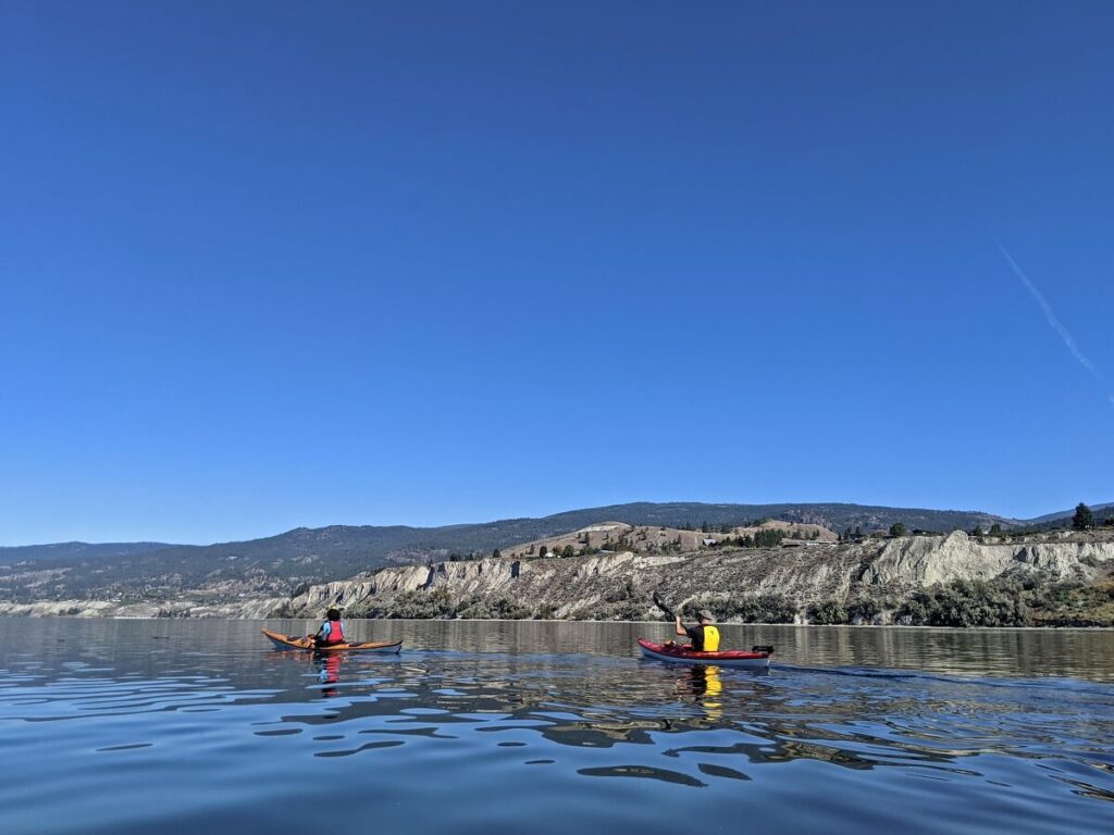 Back view of two kayakers on calm Okanagan Lake, which is bordered by tall sand cliffs. There is a bare summit above the cliffs, which is Munson Mountain